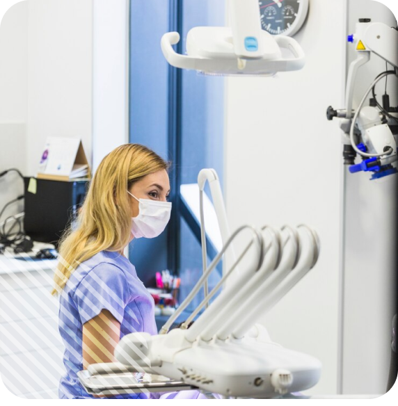 Dentist wearing a mask and scrubs in a modern dental clinic, seated beside a dental chair with various tools.